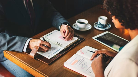 Two people facing each other at a desk with notebooks on the table