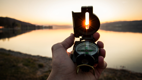 Hand holding a compass pointing towards the sunset on the horizon beside a lake