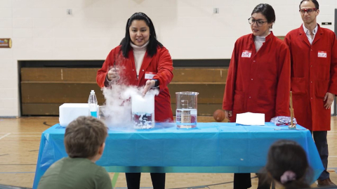 Three scientists wearing red lab coats behind a table, showing youth a bubbly experiment.