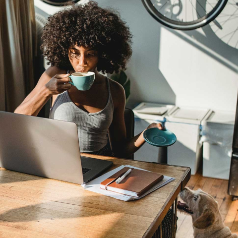 Woman sits at a desk drinking coffee working on a laptop