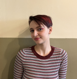 A portrait of a person with medium-length, dark-red dyed hair standing against a beige wall.