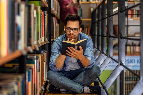 A man sits on the floor in the library, reading a book