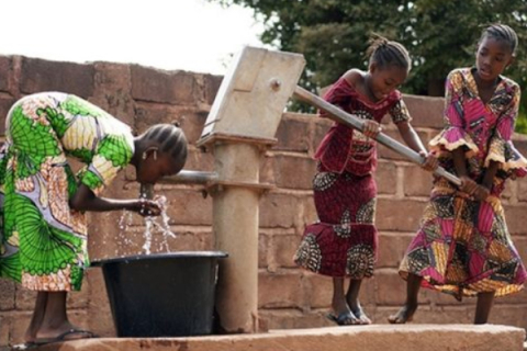 One young girl drinking from a public well while two other girls pump water