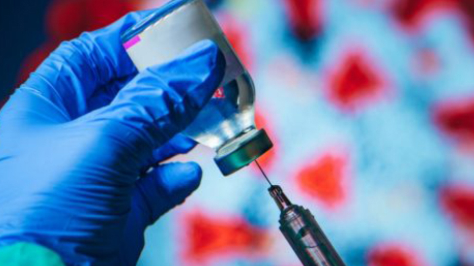 Gloved hand holding a vaccine vile while a syringe is drawing a vaccine dose with a blurred representation of the coronavirus in the background