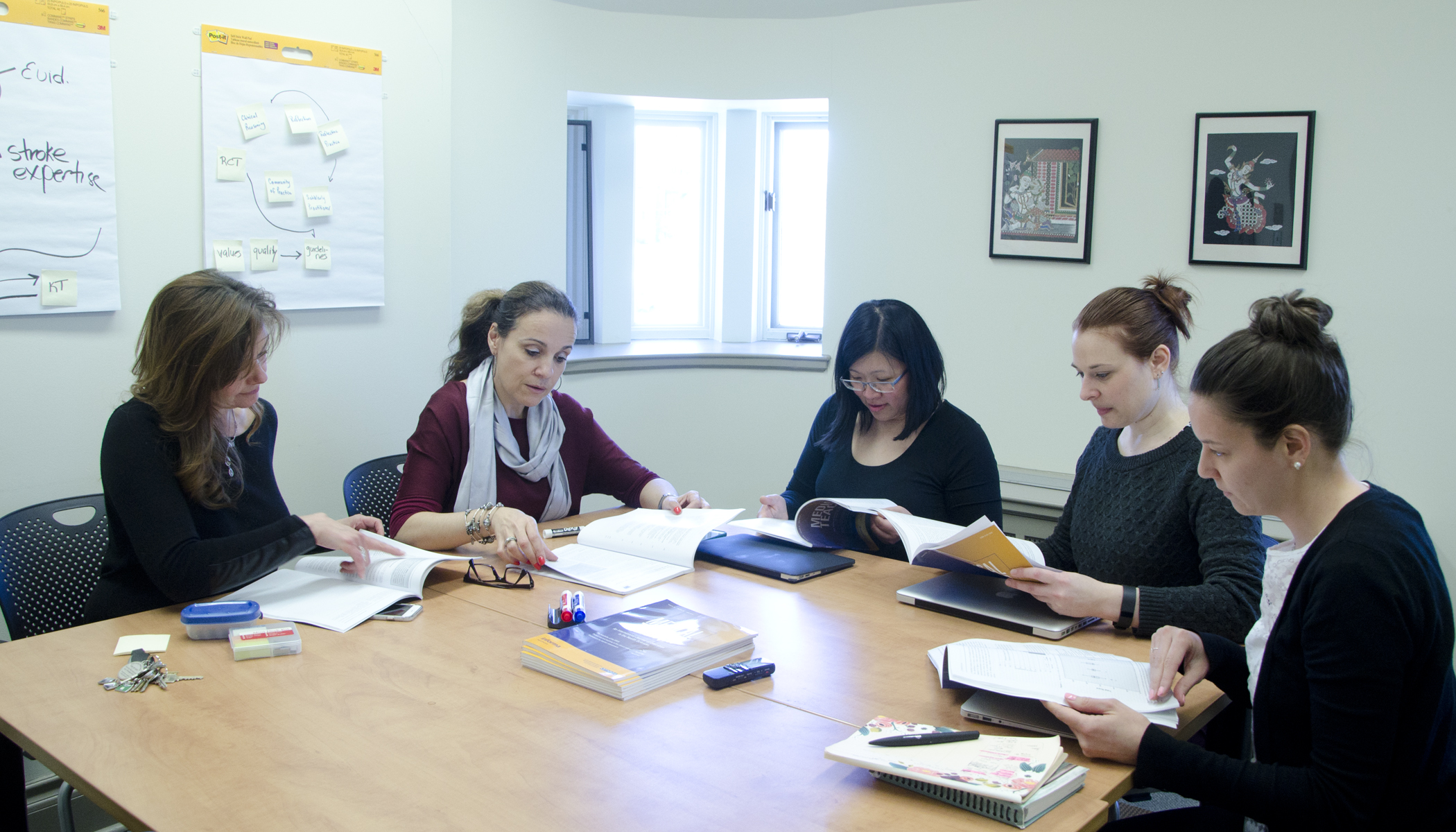 Five females sitting around a table focusing on books and devices in front of them having a conversation with each other.