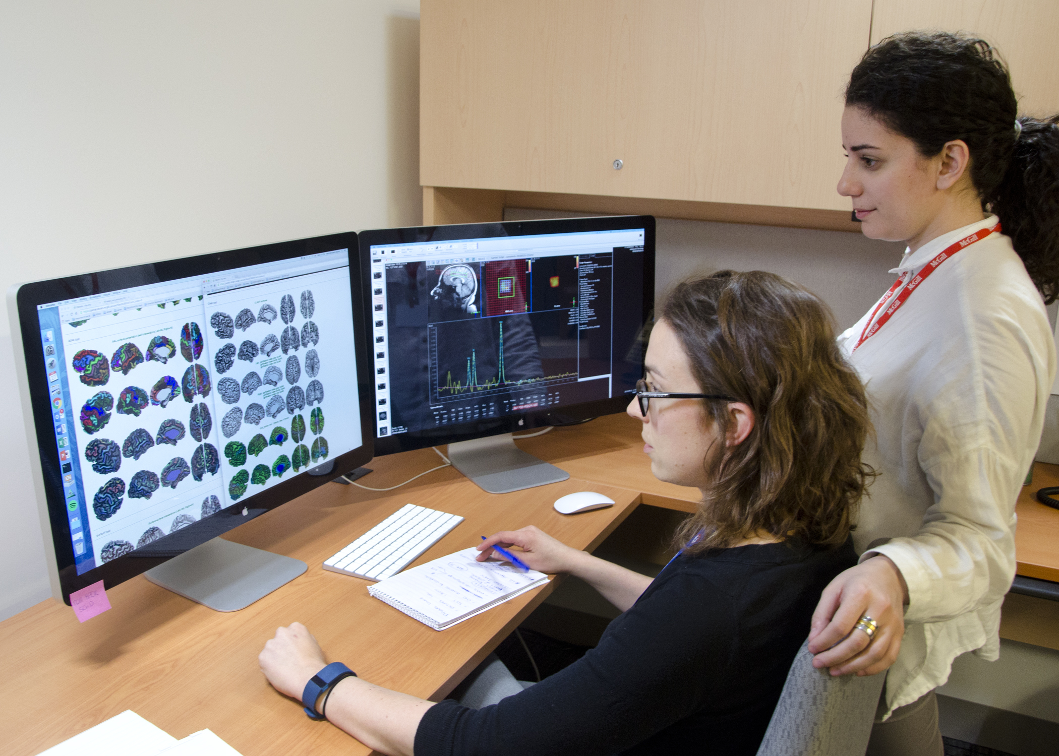 A researcher and assistant looking at brain images on two monitors.