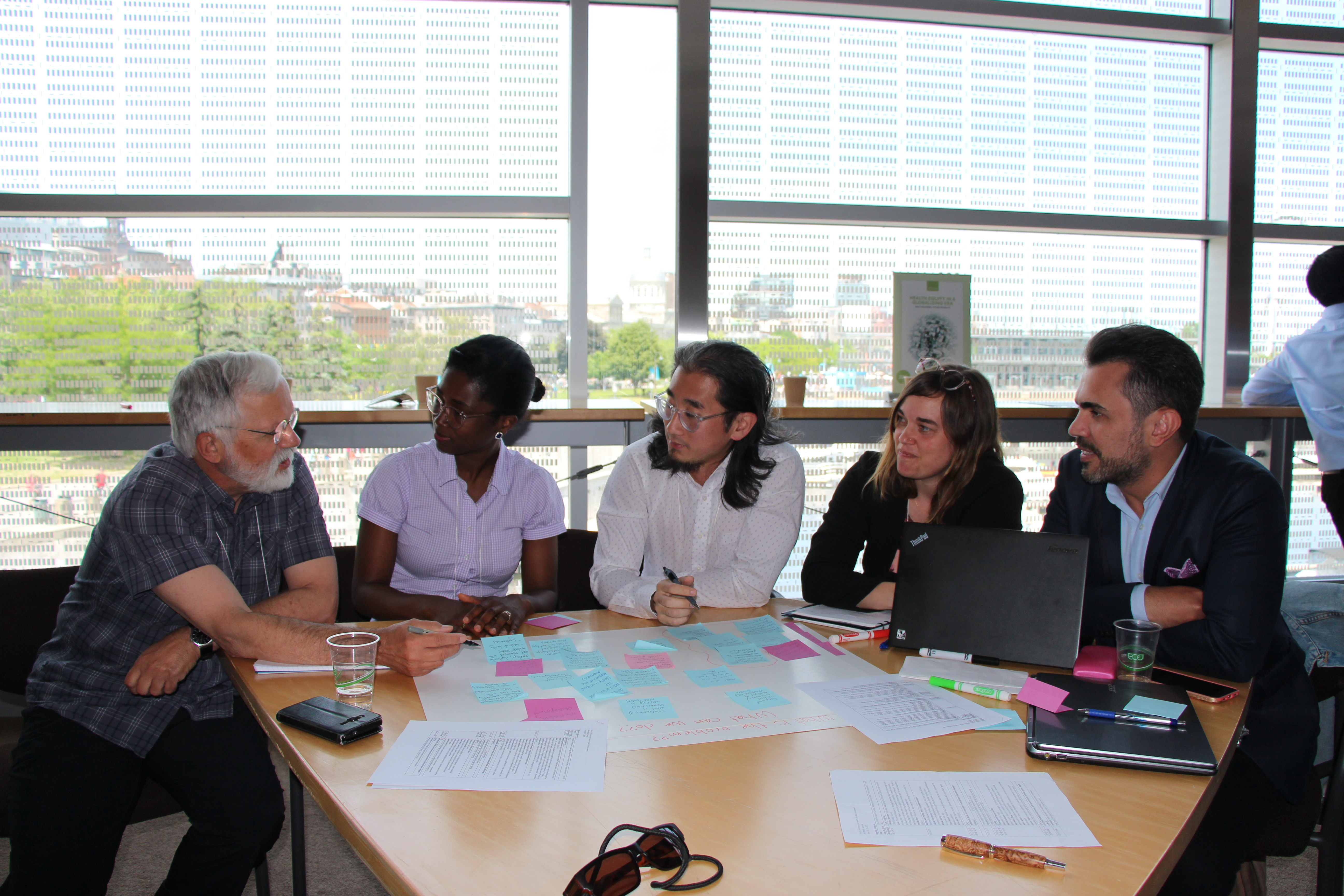 Five people, three males and two females sitting around a table discussing with papers and post it notes scattered across table. 