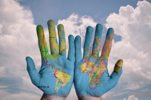 Two hands, palm side with a map of the world painted on the hands. Background is the sky. 