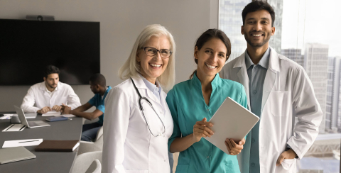 Three healthcare professionals standing together, 2 members of team in background seated at table.