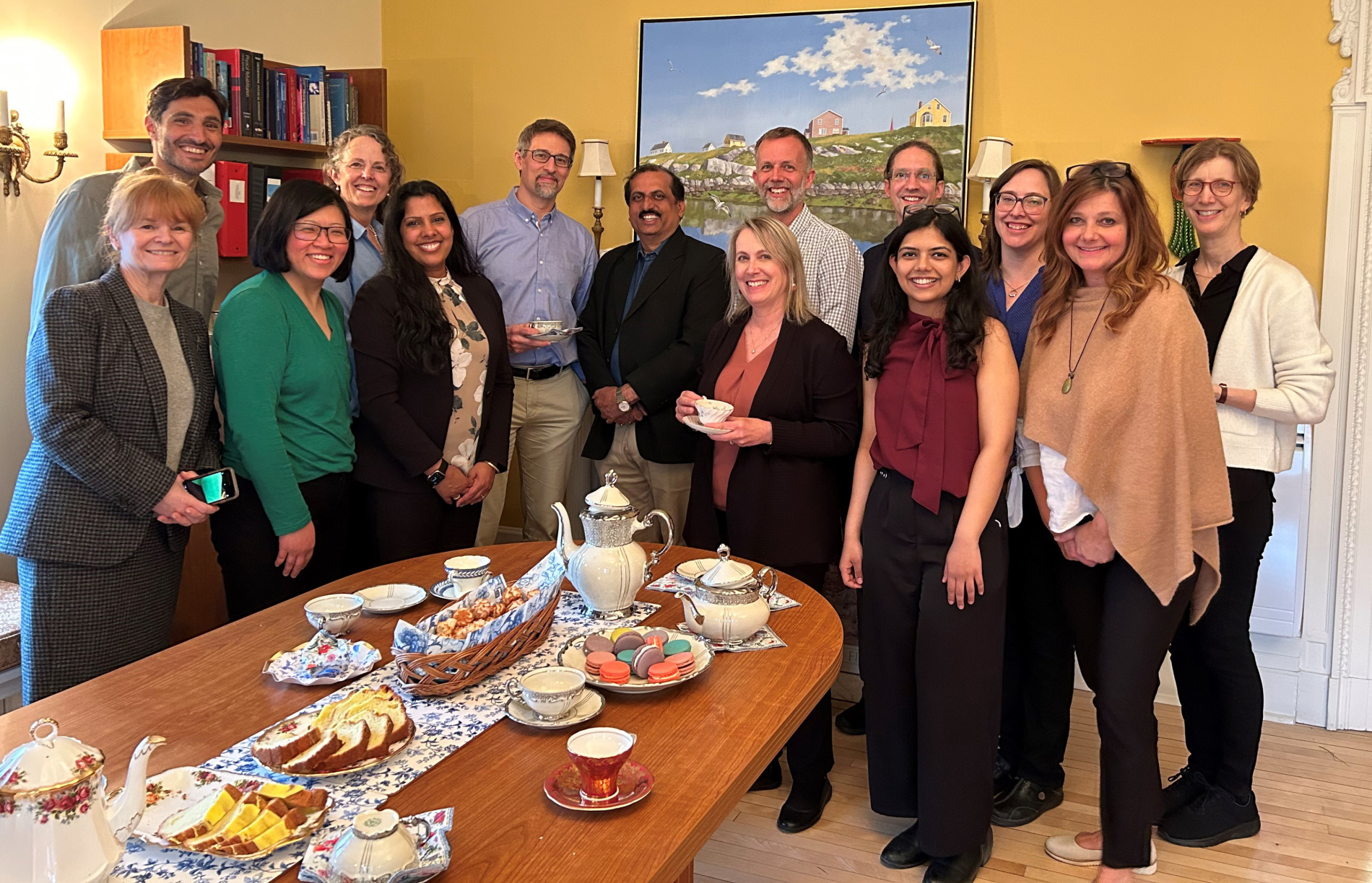 Group  photo of 14 people, in a room with tea and snacks on table, including Professor Maiya in center.