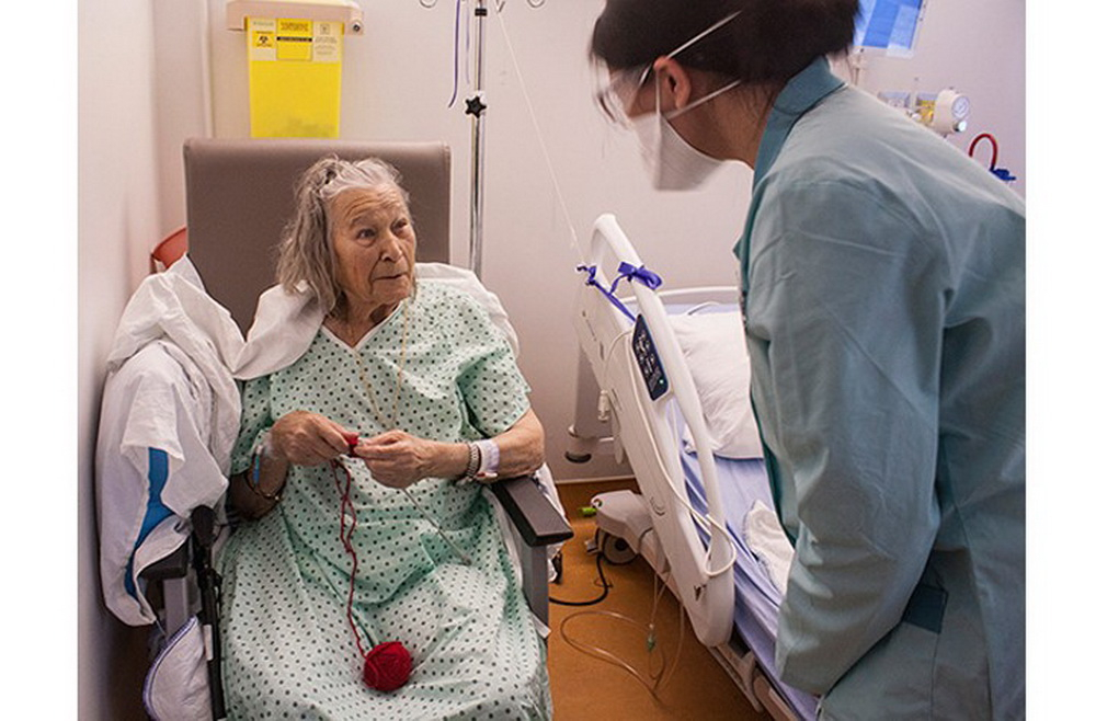 older lady patient seated and knitting while student wearing protective mask speaks to her 