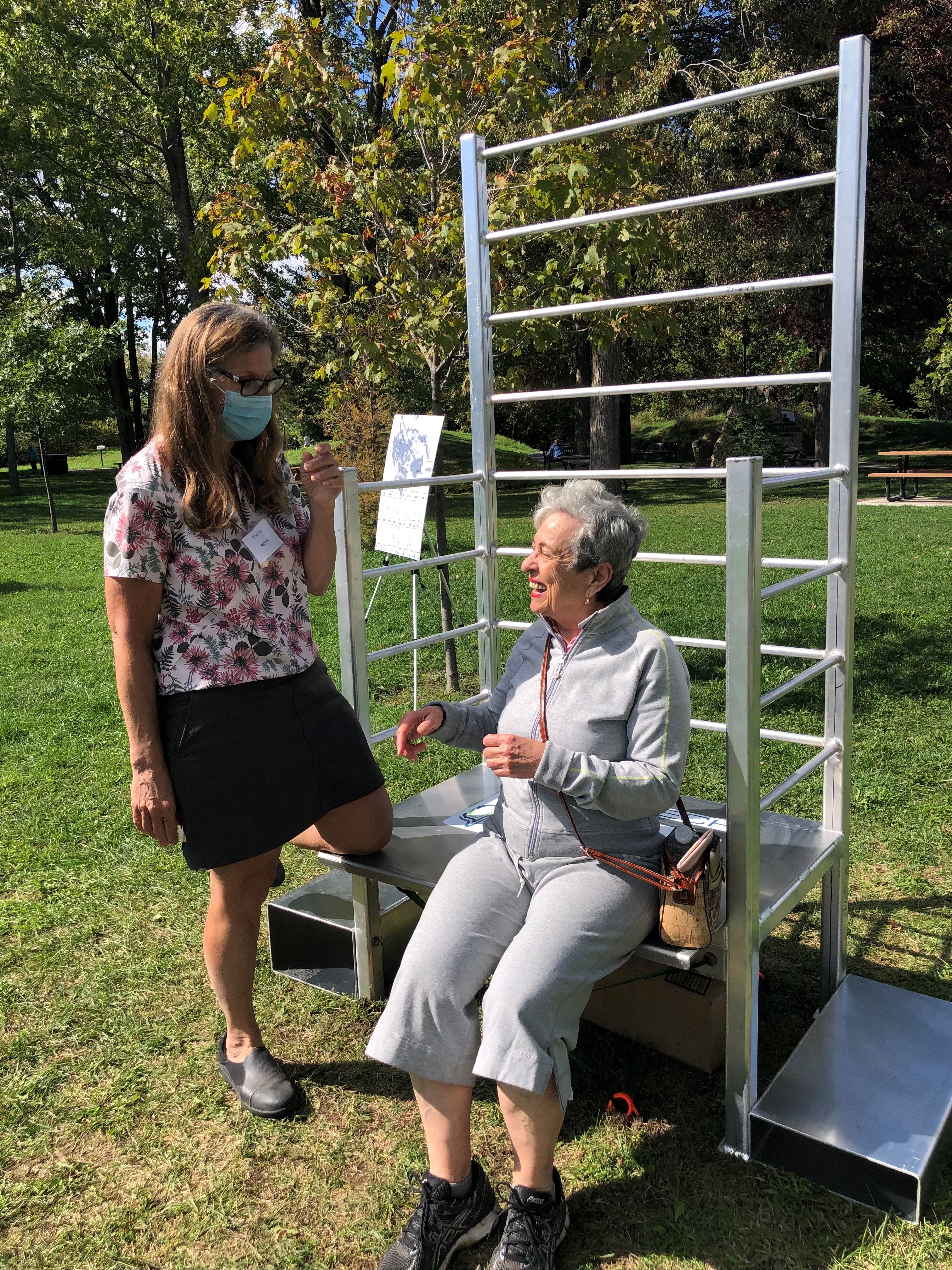 Physiotherapist explaining exercise to a women sitting on the bench part of outdoor exercise structure. 