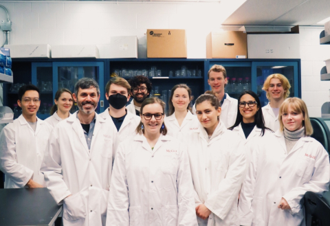 A group of students and a principle investigator wearing white lab coats smile into the camera while standing in front of lab equipment.