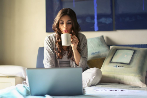 Woman sitting on bed drinking coffee looking at laptop 