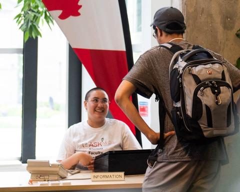 A McGill student employee, wearing glasses and an “Ask McGill” t-shirt, sits smiling at a desk while speaking with another student