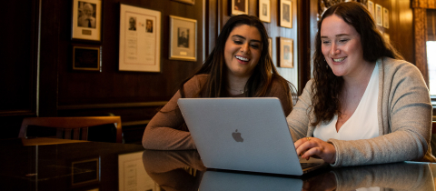2 students in a library sitting in front of a laptop smiling