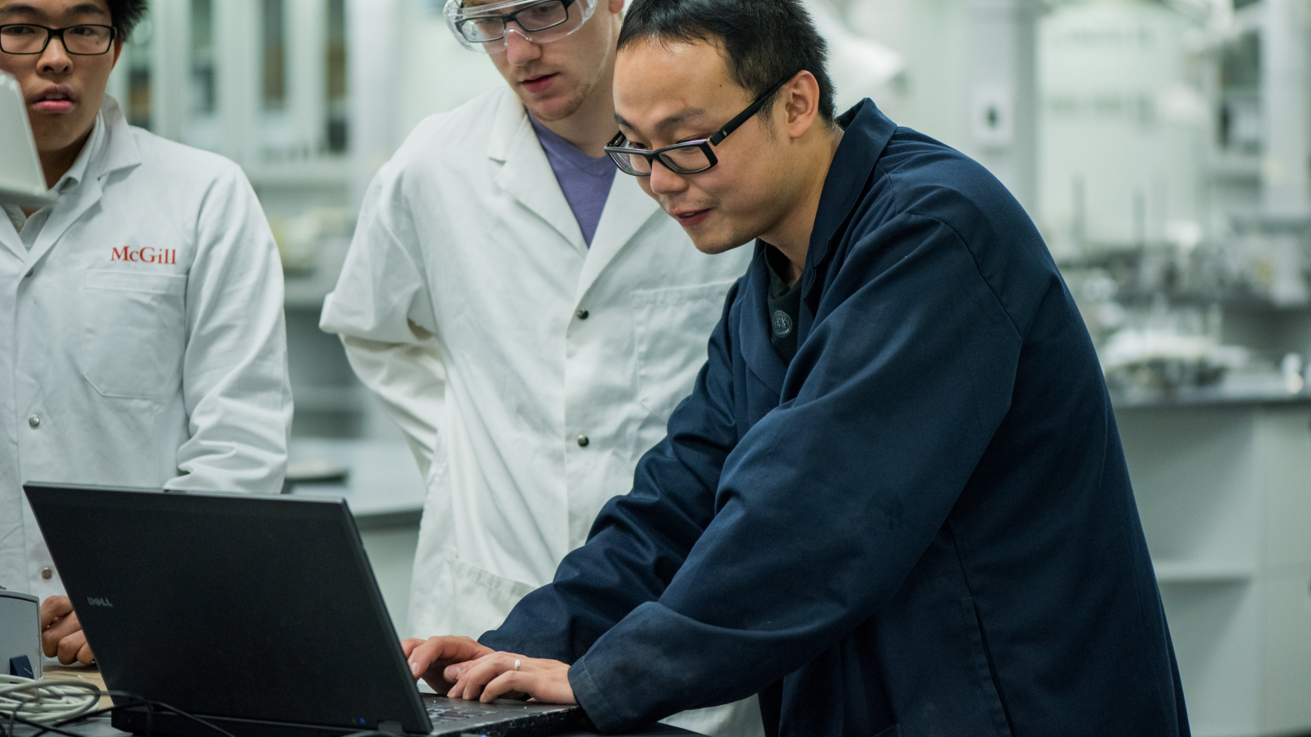 Two researchers in white McGill lab coats look on as someone types on a laptop atop a chemistry lab bench.