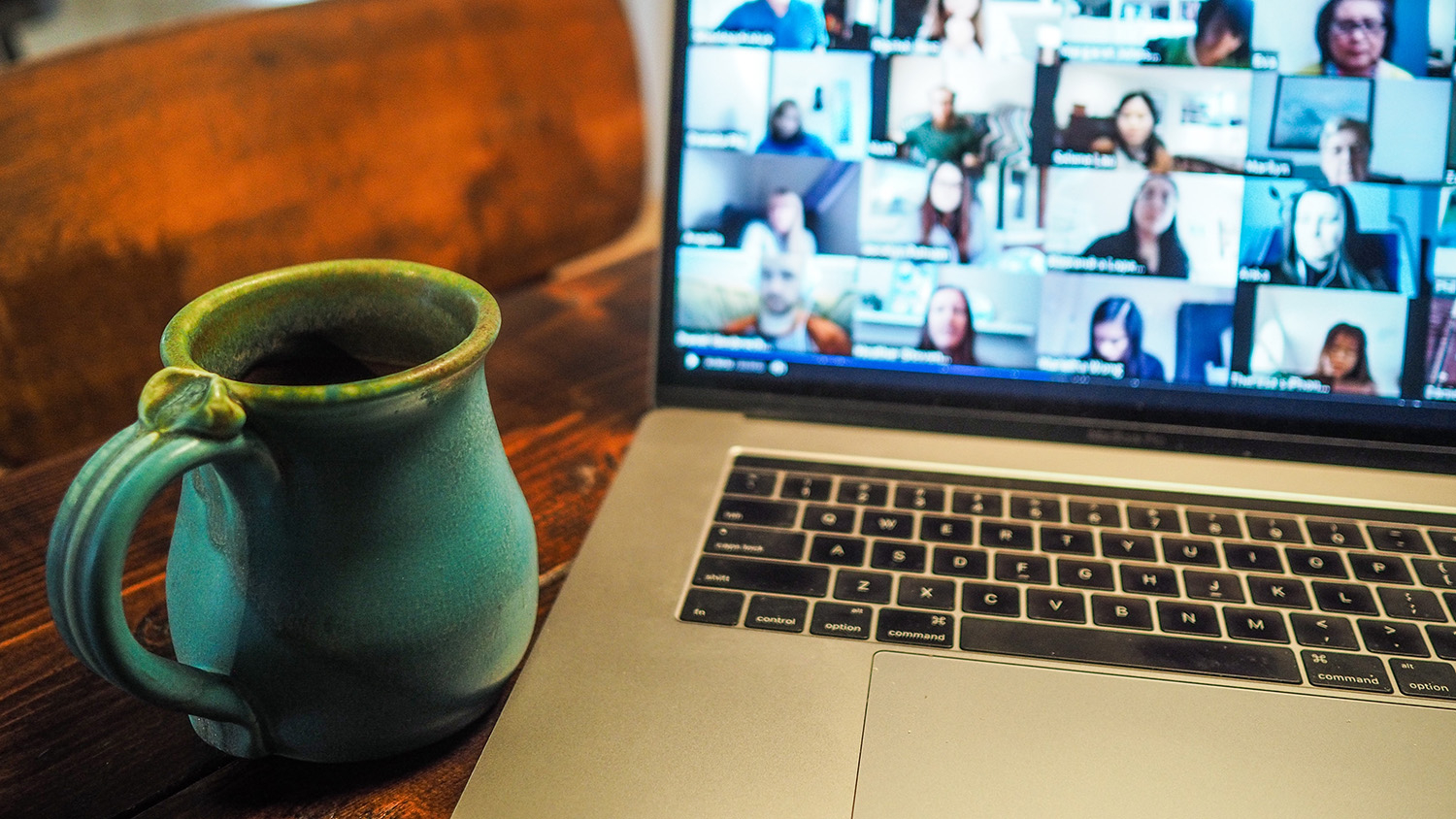 A coffee cup and laptop on a wooden desk. Numerous meeting attendees can be seen on the laptop display screen.