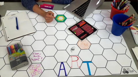 A student draws with markers on a large paper that has a honeycomb pattern printed on it.