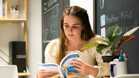 A student sits, reading a book at a table in the Healthy Living Annex, surrounded by a plant and chalkboard