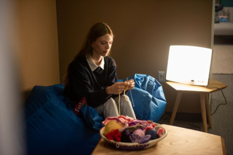 A student sits on a beanbag chair, knitting, in the lounge area of the Healthy Living Annex