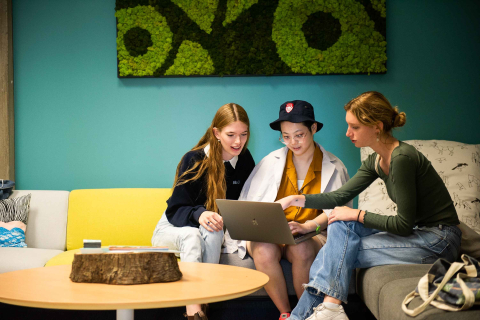 Three students sit together on a couch, looking at a laptop screen, in the Healthy Living Annex